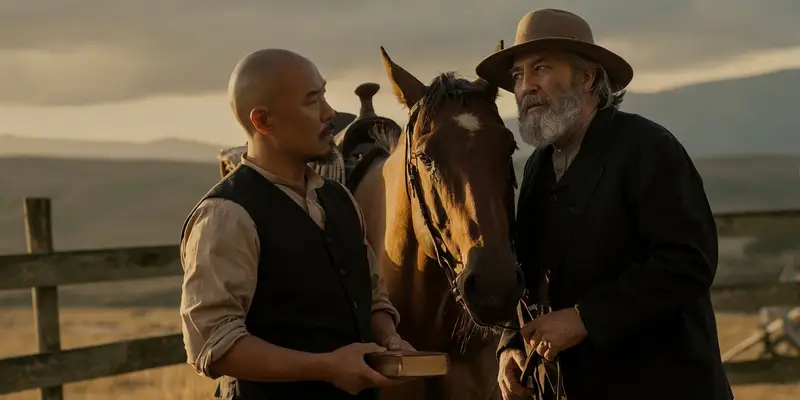 Hoon Lee as Lee, holding a book, and Ciarán Hinds as Sam Hamilton, in a wide-brimmed hat, standing either side of a horse at a ranch fence at dusk