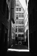 Black and white: a narrow brick alley looking towards a tall stepped-gable facade.