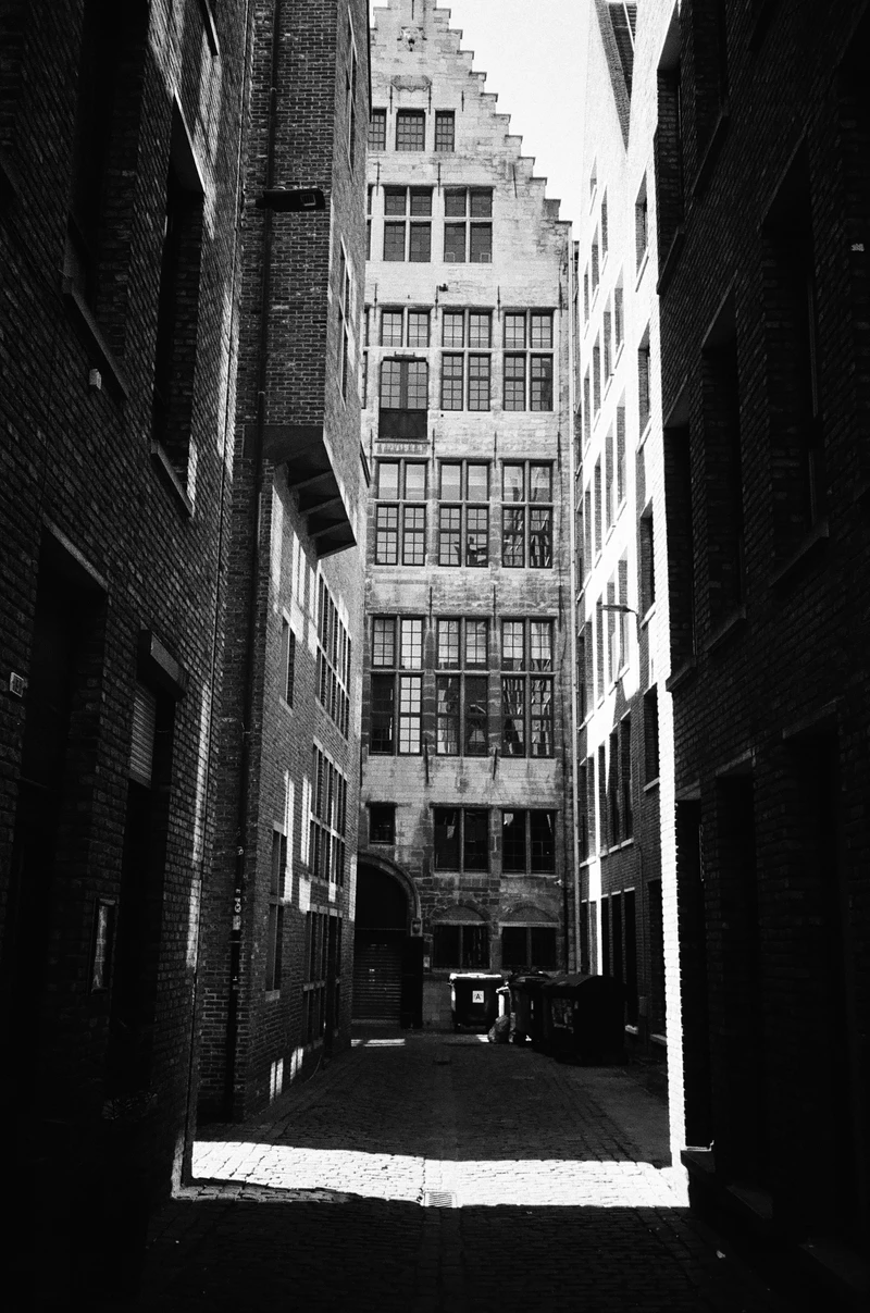Black and white: a narrow brick alley looking towards a tall stepped-gable facade.