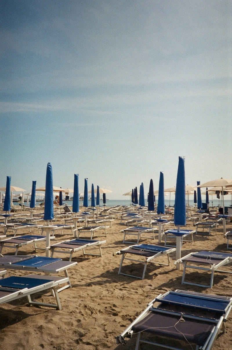 Rows of empty sunbeds and furled blue umbrellas on a sandy beach, the sea a thin line behind.