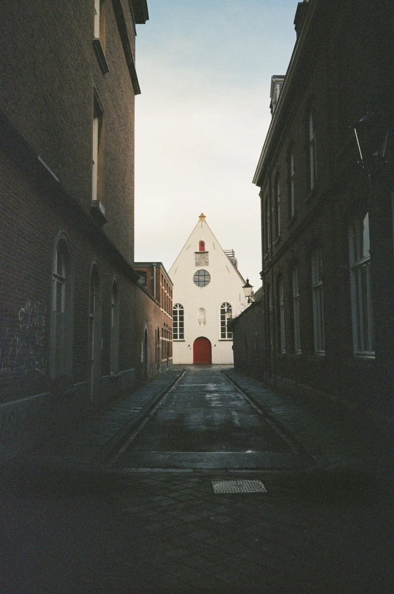 A narrow lane between dark brick houses, opening onto a white gabled chapel with a red door.