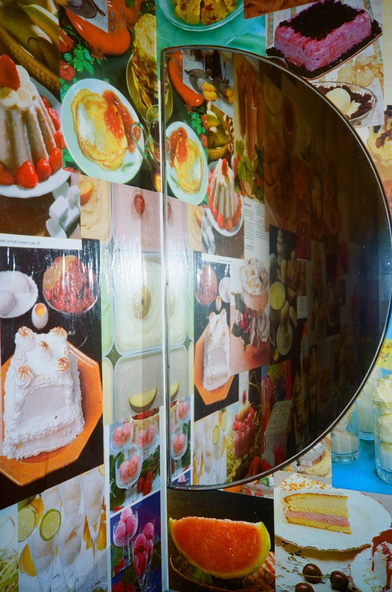 A wall papered edge to edge with cut-out pictures of cakes, jellies and fruit, a round mirror doubling the collage.