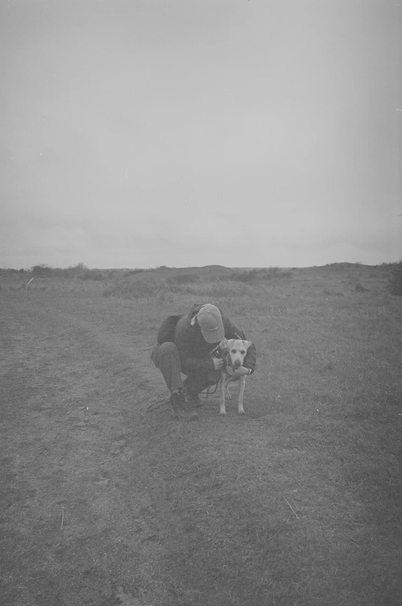 Black and white: someone crouching in dune grassland to hold a pale dog on a lead, low dunes and flat sky behind.