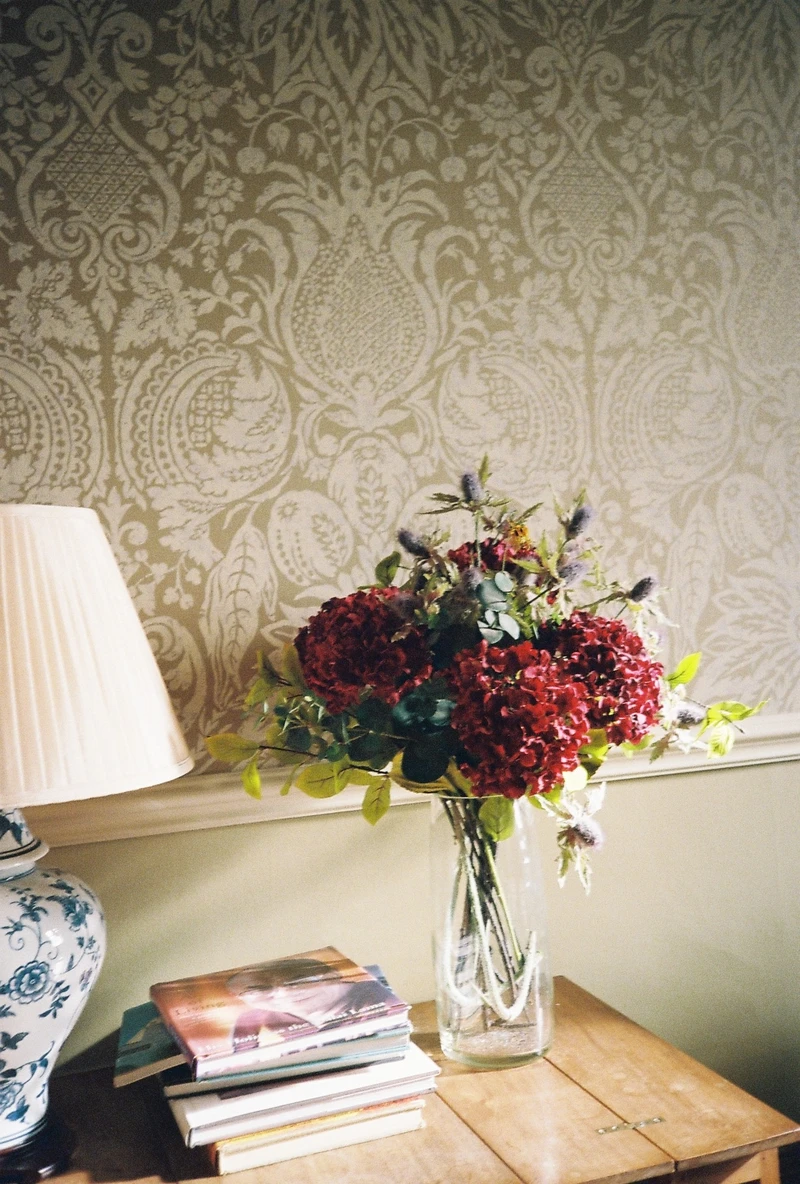 A vase of deep red hydrangeas and thistles beside a lamp and a stack of books, against damask wallpaper.