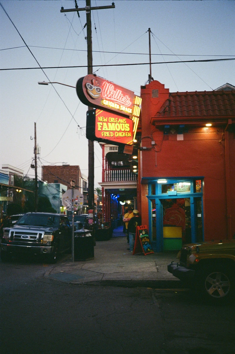A neon fried chicken sign lit at dusk on the corner of an orange building with a blue doorway, power lines overhead.
