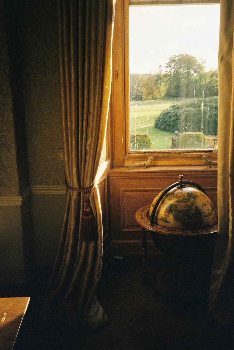 An old globe by a sash window with gold curtains, clipped shrubs and parkland outside in low sun.