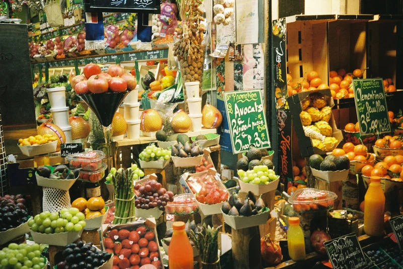 A greengrocer's display piled with grapes, figs, clementines and avocados, prices chalked on green boards.