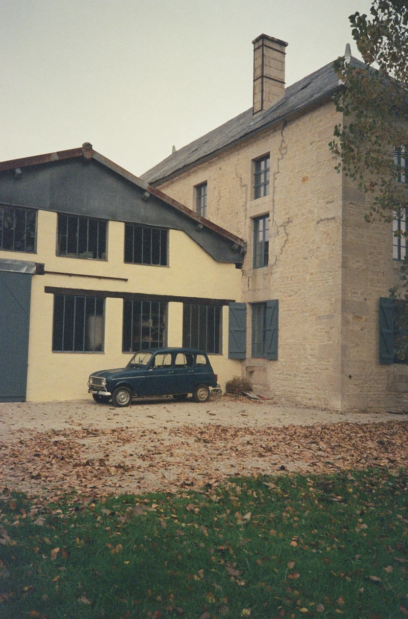 A Renault 4 parked on gravel against the side of a stone and yellow-rendered house.