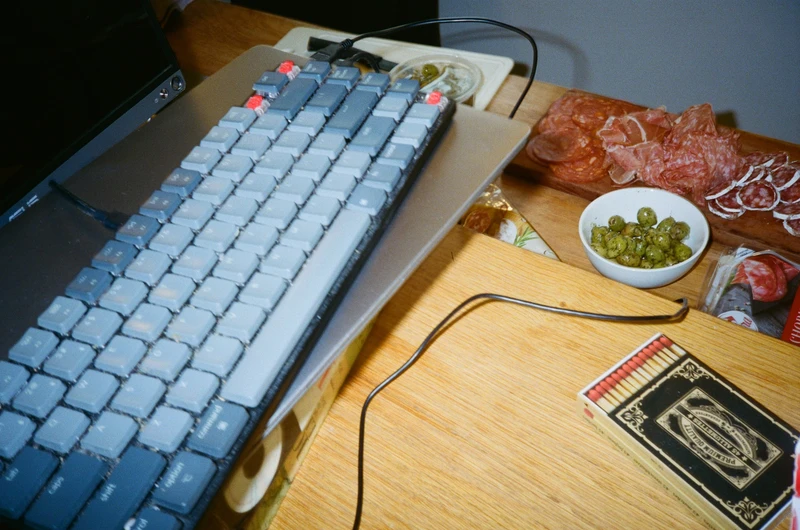 A mechanical keyboard on a laptop beside a board of cured meats, a bowl of olives and a box of matches.