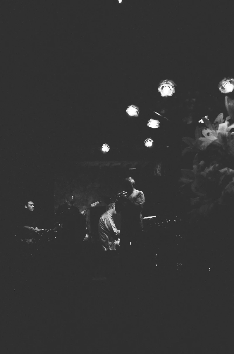 Black and white: figures at night under a scatter of blown-out lights, most of the frame in darkness.