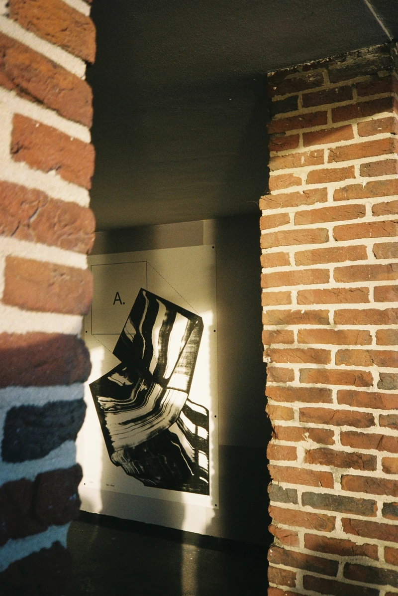 Seen between two brick walls, a poster with a black brushstroke and the letter A, lit by a slice of evening sun.