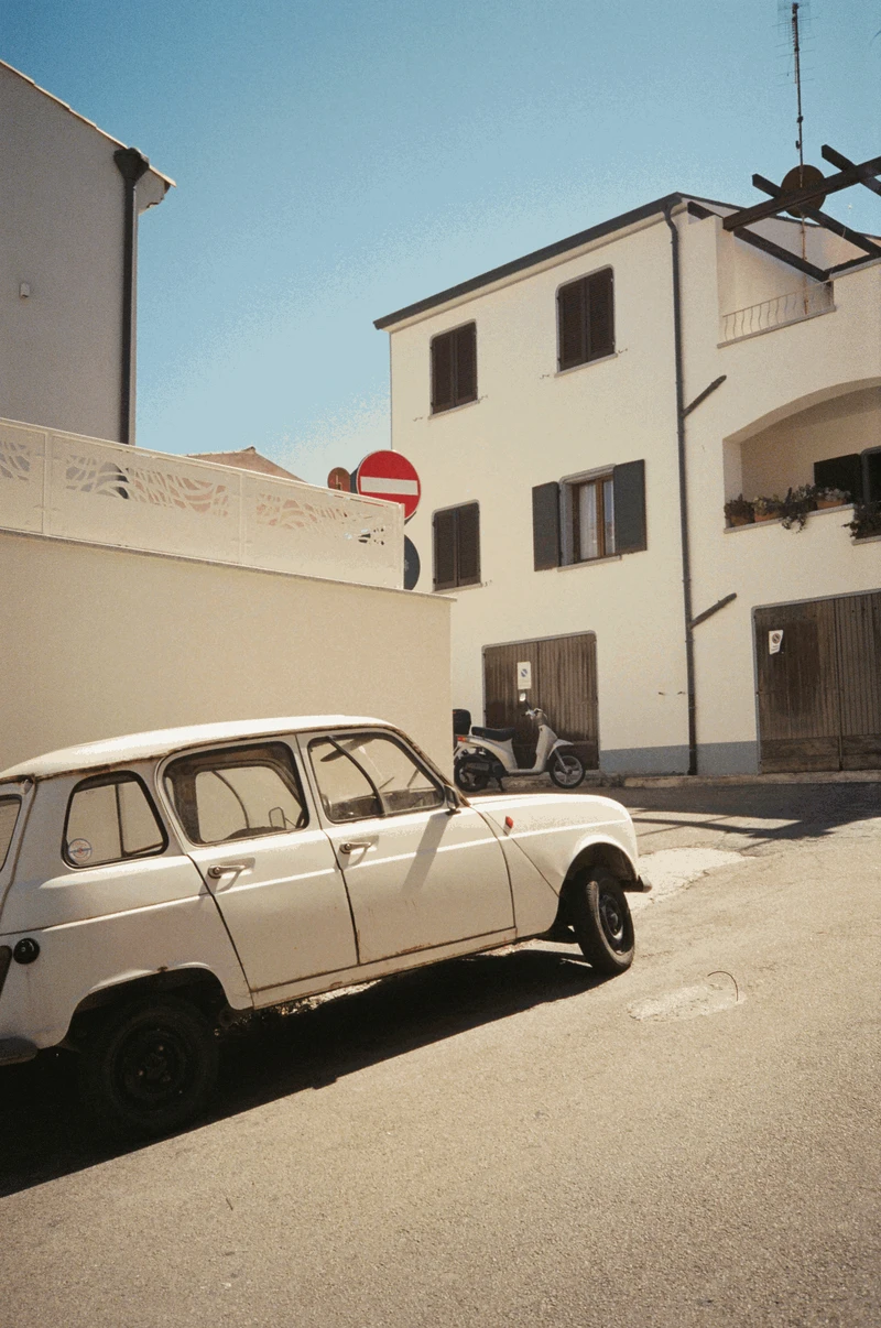 A white Renault 4 parked on a sunlit corner below a no-entry sign, a scooter and shuttered houses behind.