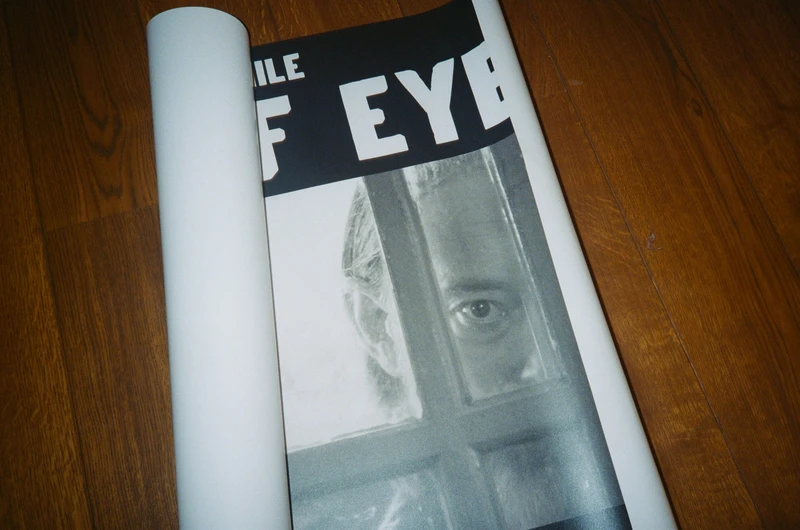 A half-unrolled poster on a wooden floor: a black and white face looking through a window pane under bold lettering.