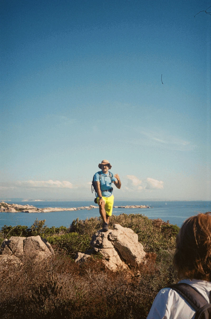 A guide in a sun hat and yellow shorts standing on a granite boulder above the sea, mid-sentence.