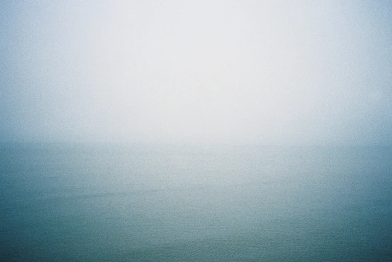 A calm sea fading into fog, the horizon lost between grey-green water and white sky.