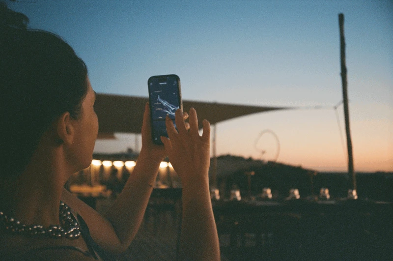 Seen over her shoulder, someone holding up a phone showing a star map against a blue and orange dusk sky.