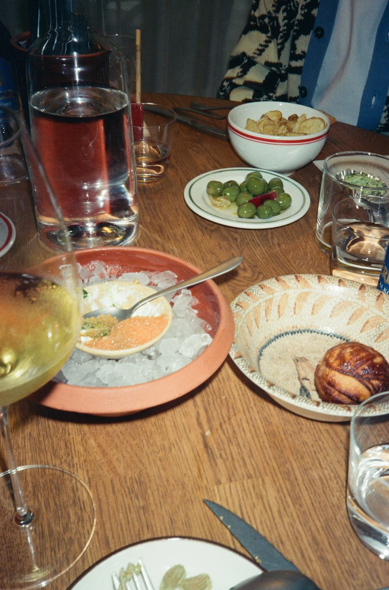 A wooden table mid-meal: olives, crisps, bread, a dish of ice, and half-drunk glasses.