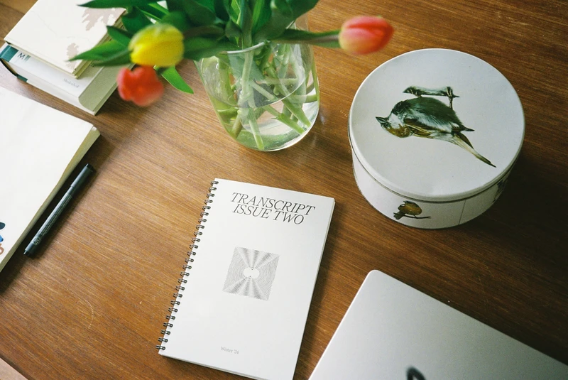 A spiral-bound copy of TRANSCRIPT Issue Two on a wooden desk, beside tulips in a glass and a bird-painted tin.