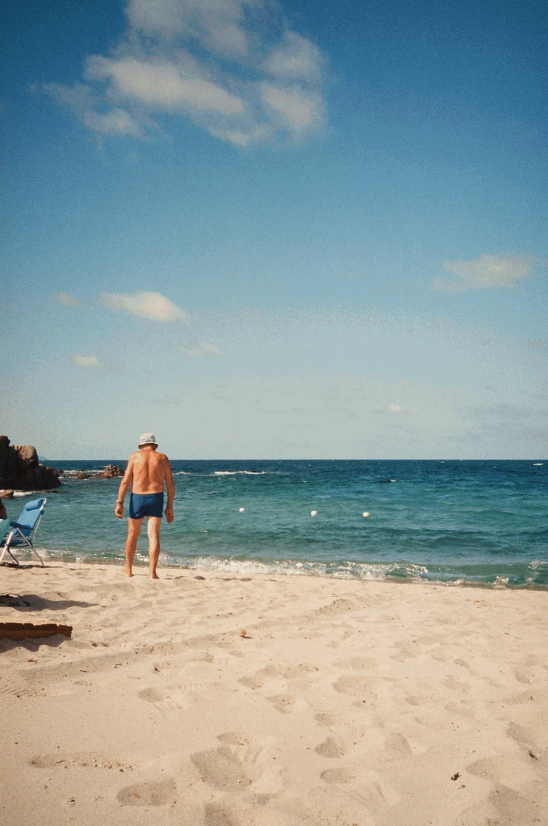 A man in a bucket hat walking down the sand towards a turquoise sea, granite rocks at the left edge of the bay.