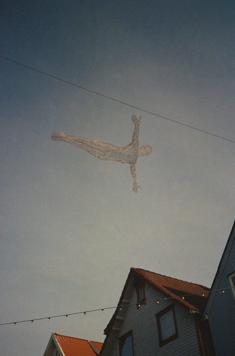 A wire figure suspended from a cable above a street at dusk, arms outstretched.